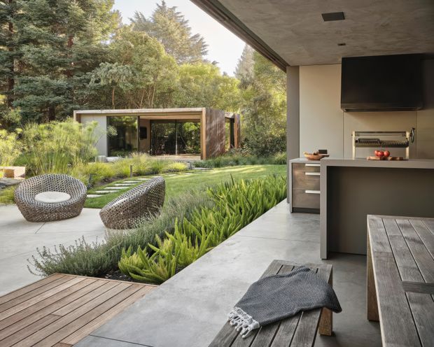 A contemporary outdoor entertaining area opening onto a landscaped garden, with a built-in barbecue kitchen, concrete flooring, timber dining table, woven lounge chairs, and a separate glass-walled pavilion among mature trees. The covered space has a broad concrete soffit and integrated television, creating a relaxed indoor-outdoor connection.