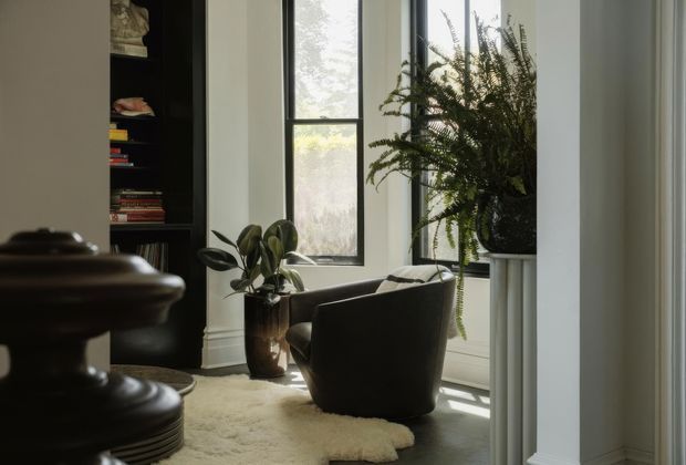 A quiet, refined reading nook with a dark upholstered armchair positioned beside tall black-framed windows. A dark built-in bookshelf filled with books occupies the left side, while potted plants add greenery near the windows and a white architectural column frames the right edge. A soft cream rug sits on dark timber flooring, creating a warm, intimate interior with abundant filtered natural light.