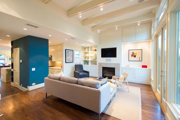 Bright open-plan living room with a contemporary-classic fit-out. A neutral sectional sofa faces a fireplace and wall-mounted television, with built-in white cabinetry and panelled walls. The room connects directly to a kitchen area, features exposed painted ceiling beams, dark timber flooring, large glazed doors or windows, and a deep blue accent wall.