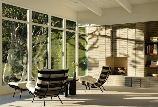 Sunlit mid-century-inspired living room with expansive floor-to-ceiling glazing overlooking mature greenery. The room features a pale brick feature wall with a built-in fireplace and open shelving, a white painted beamed ceiling, light carpet, and black-and-white lounge chairs arranged around a small central table.