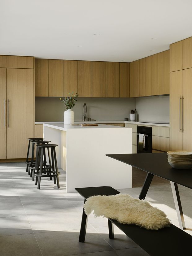 A contemporary open-plan kitchen and dining area with extensive warm timber cabinetry, a white island with integrated sink, pale tiled flooring, and a dark dining table with matching benches and stools. The space has clean, understated detailing, a neutral palette, and abundant natural light.