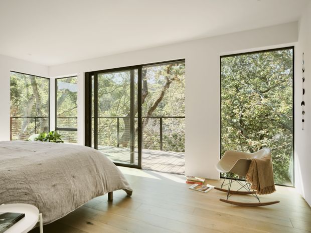 Bright contemporary bedroom with a low-profile bed, pale timber flooring, white walls and ceiling, and extensive dark-framed floor-to-ceiling glazing opening toward a leafy outdoor setting. A sculptural lounge rocking chair with a draped throw sits beside the windows, creating an airy, calm retreat.