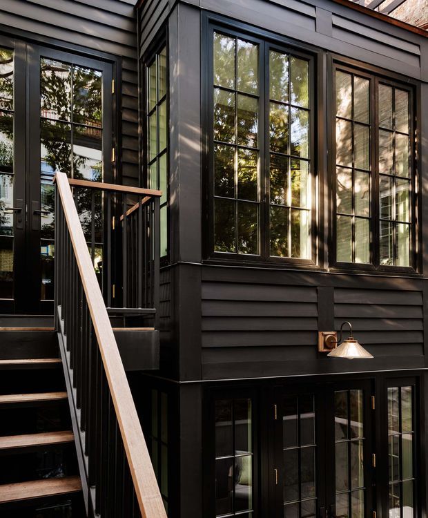 Exterior view of a dark-clad multi-level building with a stepped entry stair, black-framed divided-light windows, glazed doors, horizontal siding, and a wall-mounted exterior lamp. Trees are reflected in the glazing, while warm sunlight highlights the facade.