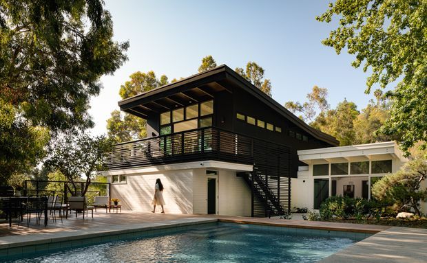 A modern two-storey residential house set among mature trees, photographed from beside a swimming pool. The dark upper volume has broad roof overhangs, extensive glazing, horizontal slatted cladding, and a metal-railed balcony. A lighter lower level opens onto a large pool terrace, with an attached white-glazed pavilion or sunroom to the right. Exterior stairs connect the balcony to the terrace.