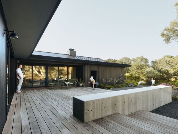 A contemporary single-storey home arranged around a spacious timber deck and concrete-edged outdoor area. Dark vertical cladding, broad glazed doors, a low-pitched metal roof, and a long raw concrete bench or retaining element define the architecture. The deck opens to landscaped surroundings with mature trees and several people present.