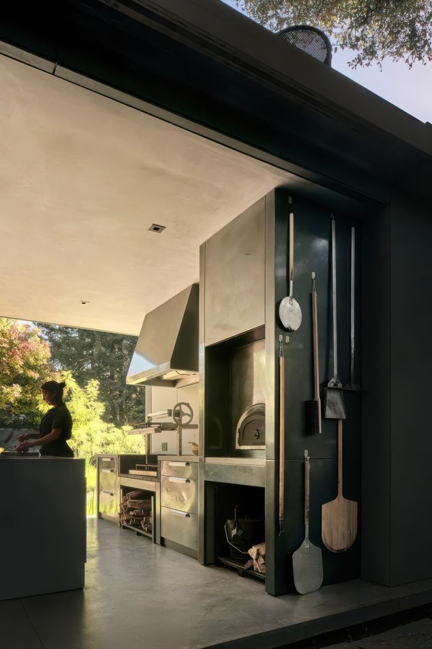 A dark, sophisticated covered outdoor kitchen with a large stainless-steel cooking and preparation area, built-in pizza oven, grill, cabinetry, and integrated storage. Cooking tools and pizza paddles are displayed vertically on a black wall panel. The space opens onto a leafy garden and features a broad pale ceiling canopy, dark metal framing, and polished concrete flooring.