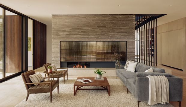 A refined contemporary living room with a large grey sectional sofa, leather lounge chairs, a timber coffee table, and a linear fireplace set into a textured horizontal stone feature wall. Full-height dark-framed glazing lines the left side, while timber-look built-in cabinetry and a dark vertical screening detail define the right side. The room has a warm neutral palette and carefully integrated architectural joinery.
