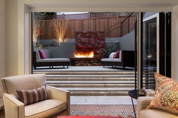 A refined indoor-outdoor living area opening onto a stepped courtyard. The foreground contains upholstered armchairs and a patterned rug, while large glazed doors frame an outdoor lounge with built-in seating, a brick fireplace, and a lit fire. Layered steps connect the interior to the courtyard, with timber fencing, masonry walls, planting, and integrated lighting creating a warm, sophisticated atmosphere.