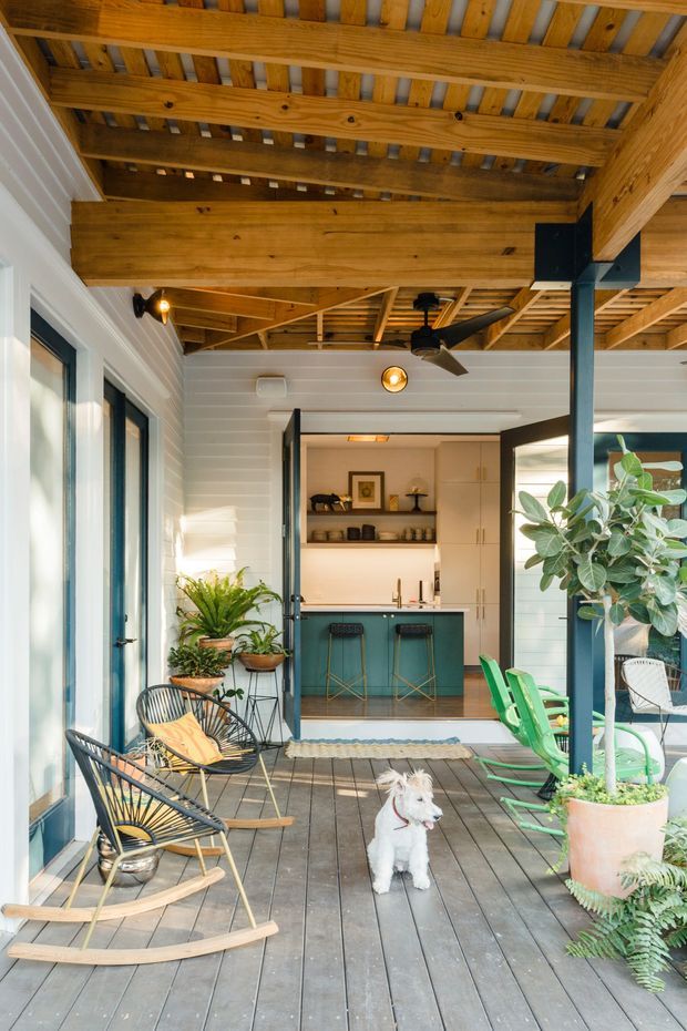 A bright covered outdoor deck or verandah with timber decking, exposed timber roof structure and beams, white horizontal wall cladding, dark-framed glazing, and a compact kitchen opening onto the space. The deck is furnished with rocking chairs and outdoor seating, surrounded by potted plants, with a small dog in the foreground. A dark steel post and ceiling fans add structural and functional details.