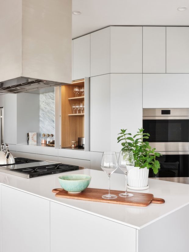 Bright contemporary kitchen with extensive matte white handleless cabinetry, integrated stainless-steel ovens, a large white island, gas cooktop, and a timber open display niche. A marble-look splashback, timber serving board, glassware, bowl, and potted plant add understated warmth. The compact vertical composition suggests a well-organised medium-sized kitchen.