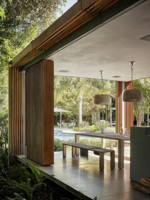 A refined open-plan indoor-outdoor dining area with a large timber-framed opening overlooking a landscaped garden and swimming pool. The space features vertical timber screening, a long timber dining table with matching bench seating, polished concrete flooring, a white plasterboard ceiling with recessed lights, and sculptural pendant lights. Lush planting and pool views create a warm, tropical connection to the outdoors.