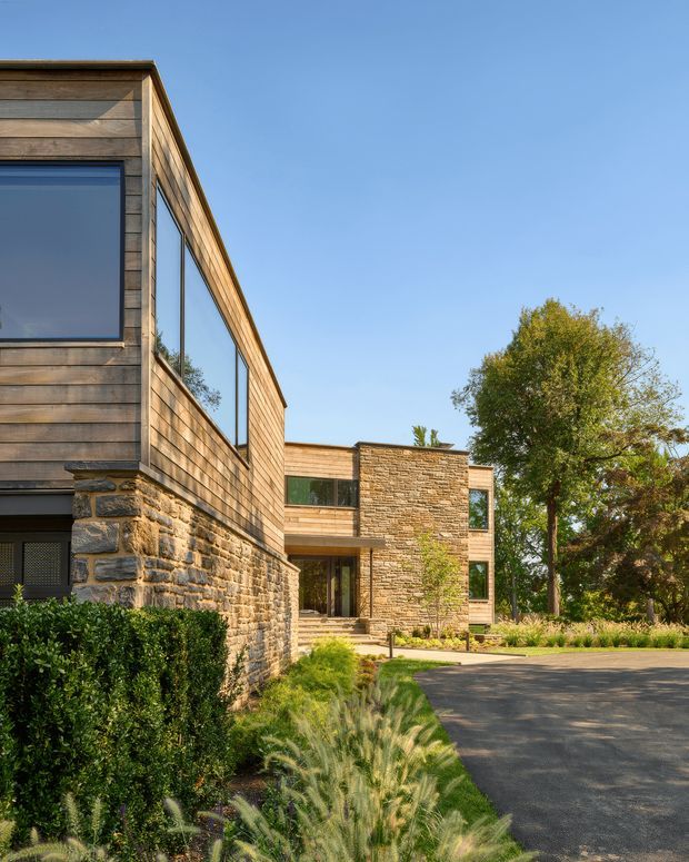 Exterior view of a contemporary low-rise residence composed of warm horizontal timber cladding, large dark-framed windows, and substantial rough natural-stone masonry walls. The landscaped approach includes clipped hedges, ornamental grasses, paving, and mature trees under a clear blue sky.