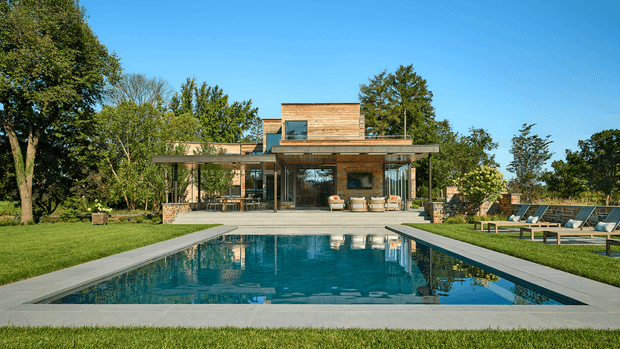 A modern flat-roofed house set within a landscaped garden, viewed from across a rectangular swimming pool. The home features extensive floor-to-ceiling glazing, projecting roof planes, warm timber cladding, and a broad covered outdoor living area. Poolside loungers and paving frame the foreground, with mature trees and lawn surrounding the property.