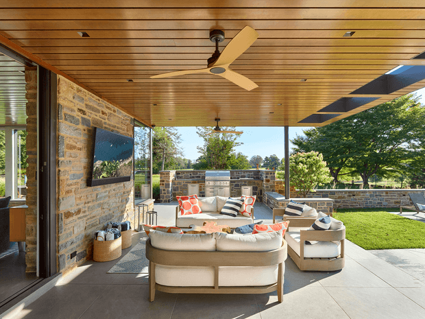 A spacious covered outdoor living area connected to a stone-clad home, featuring a timber-lined ceiling with an integrated ceiling fan, large-format paved flooring, built-in outdoor kitchen cabinetry and barbecue, a television mounted on the stone wall, and contemporary upholstered lounge seating. The open sides overlook a landscaped lawn and garden, creating a bright, refined indoor-outdoor entertaining space.