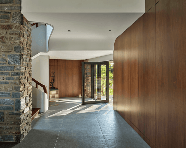 A refined residential entry hall with a broad grey stone-tile floor, full-height dark timber wall panelling, and a contrasting exposed natural-stone structural wall. A curved white ceiling and sculptural stair enclosure lead toward a glazed double entry door, which opens to greenery outside. Strong daylight casts long shadows across the floor, highlighting the clean contemporary detailing and warm material palette.