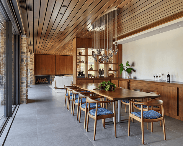 Open-plan dining and living area with a long timber dining table surrounded by upholstered wooden chairs, a timber-lined ceiling, extensive built-in timber cabinetry, stone masonry columns, and a sculptural hanging light. Large glazing opens along the left side, while a lounge and fireplace are visible in the background. The space has a refined, warm contemporary character with natural materials and a strong indoor-outdoor connection.