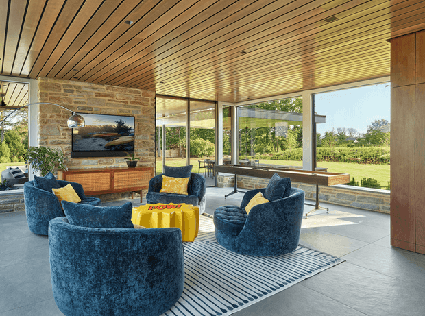 A bright, expansive contemporary living room with floor-to-ceiling glazing overlooking a landscaped garden. The room features a striking natural stone fireplace wall with a television, warm timber slatted ceiling, large grey floor tiles, blue sculptural lounge chairs, yellow accent cushions and ottomans, and a long timber console beneath the windows. The space has strong indoor-outdoor visual connection and abundant natural light.
