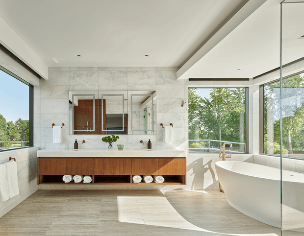 Bright, spacious contemporary bathroom with a freestanding white bathtub beside full-height glazing, a floating dark timber double vanity with open towel storage, large pale stone-look wall surfaces, and a frameless glass shower enclosure. Natural light and views of leafy trees create an airy, refined atmosphere.