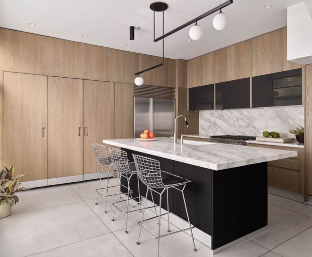 Contemporary kitchen with full-height light timber cabinetry, a large black island, and a thick white marble-look benchtop. Three wire-frame bar stools sit at the island, while integrated stainless-steel refrigeration, dark upper cabinetry, a gas cooktop, and modern pendant and track lighting complete the clean, refined fit-out.