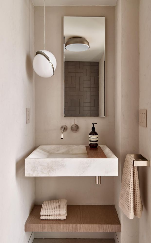 A narrow, refined powder room with a floating white stone wall-mounted basin, brushed metal tapware, a tall rectangular mirror, and a geometric textured feature wall visible in the reflection. Warm beige plaster walls, integrated spherical wall lighting, timber-toned lower shelving, and neatly arranged neutral towels create a calm, minimalist interior.