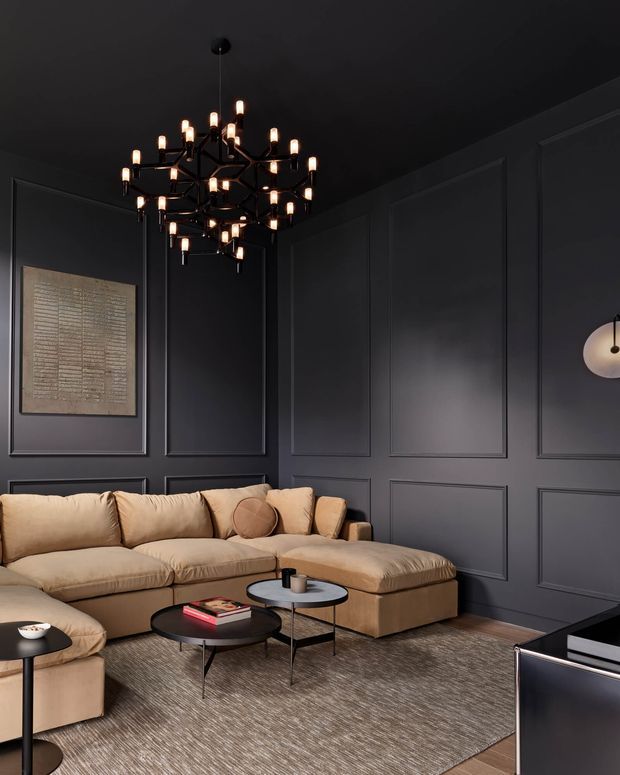A moody, luxurious living room with charcoal-grey wall panelling and ceiling, a large tan sectional sofa, layered round coffee tables, a textured neutral rug, and a dramatic black multi-arm chandelier. A framed artwork and sculptural wall sconce add restrained decorative detail, while a television is visible at the lower right.