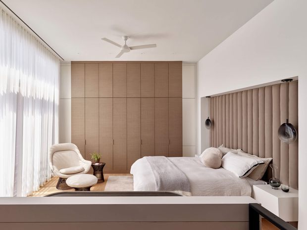 Bright, refined bedroom with a neutral palette and floor-to-ceiling sheer curtains. The room features a large upholstered bed with a vertically channelled padded headboard, full-height timber-toned built-in wardrobes, minimalist bedside tables and pendant lights, a sculptural lounge chair, and a ceiling fan. The design is calm and contemporary with warm natural finishes.