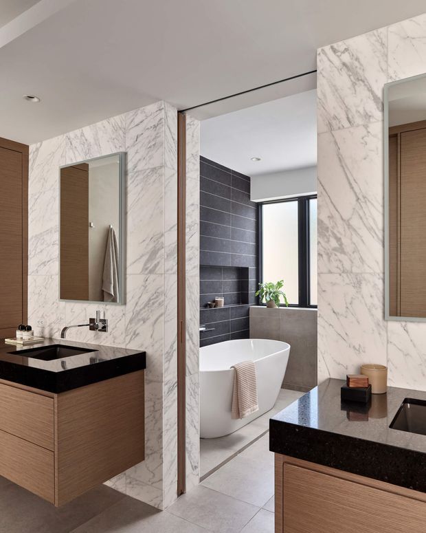 Contemporary bathroom with a double-vanity arrangement, black stone benchtops, warm timber cabinetry, and dramatic white marble-look wall cladding. A freestanding white bathtub is visible through the open partition, with dark charcoal wall tiles, a recessed niche, and a tall window providing natural light. Large mirrors and clean recessed lighting create a refined, high-contrast interior.