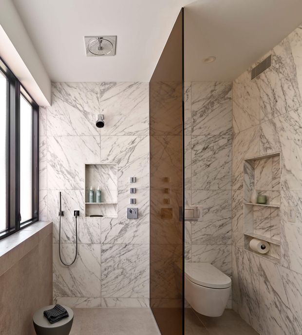 Contemporary bathroom with a marble-look wall and floor finish, frameless glass shower enclosure, wall-mounted shower fixtures, recessed storage niches, wall-hung toilet, and a large black-framed window. The space has a clean, refined appearance with light neutral surfaces and dark accents.