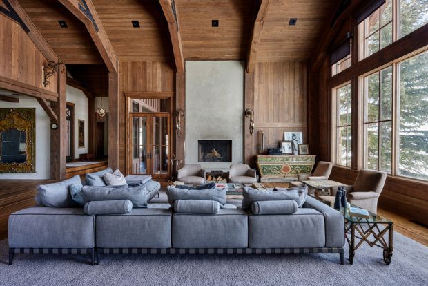 A large, rustic yet refined living room with exposed timber beams, extensive vertical timber wall and ceiling panelling, tall divided timber-framed windows, and a central plastered fireplace wall. A substantial grey sectional sofa faces the fireplace, with leather and upholstered armchairs, a decorative antique-style console, and layered traditional furnishings. The room has a high timber ceiling, strong natural light, and views of trees through the windows.