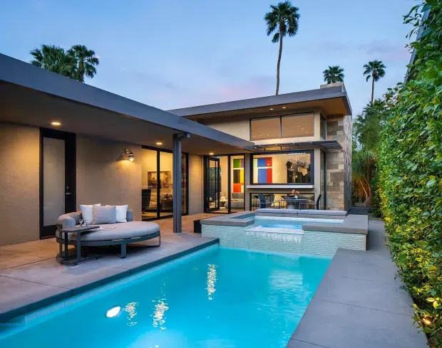 Twilight exterior view of a modern single-storey courtyard residence with flat roofs, extensive glazing, dark rendered walls, stone-clad sections, and a covered outdoor lounge beside a rectangular swimming pool and raised spa. Tropical palms and dense screening vegetation surround the property.