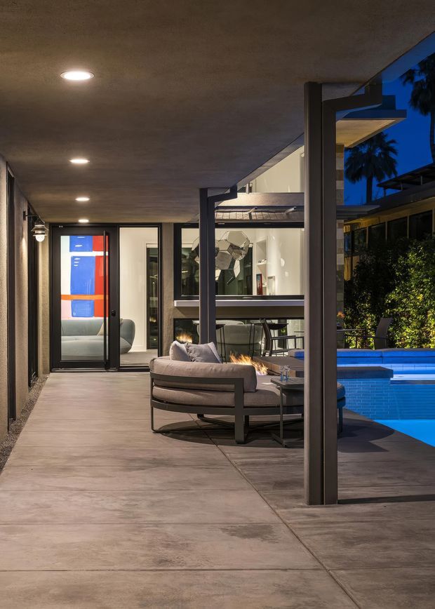 A dusk view of a covered outdoor living terrace beside a swimming pool. The space features a broad concrete-paved floor and ceiling, dark structural posts and framing, recessed downlights, a cushioned outdoor lounge setting, and a linear outdoor fire feature. Glazing opens onto a brightly lit contemporary interior, while the pool and landscaped courtyard are visible to the right.