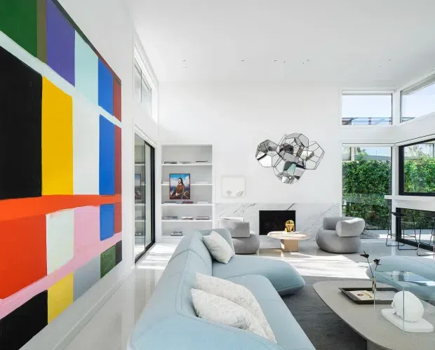Bright contemporary living room with a sculptural pale blue sectional sofa, rounded grey armchairs, a glass coffee table, white built-in shelving, expansive black-framed windows, and a large colourful geometric abstract wall feature. The room has a high white ceiling and abundant natural light.