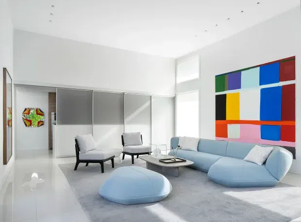 Bright, spacious contemporary living room with a pale blue curved sectional sofa, matching ottoman, two black-framed lounge chairs, and a low glass-topped coffee table. The room has glossy white flooring, white walls and ceiling with recessed downlights, translucent sliding panels or doors, tall glazing, and a large colourful geometric artwork as a focal point.