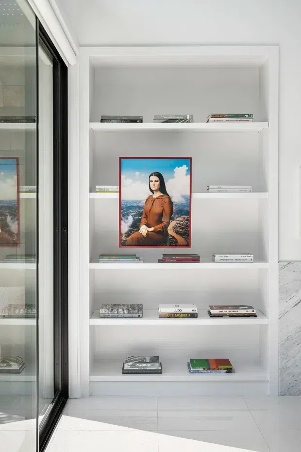 Bright, minimalist built-in white shelving displaying art books and a framed portrait, adjacent to a black-framed glazed opening or cabinet. White tiled flooring and a partial marble-look wall surface reinforce the clean contemporary interior.