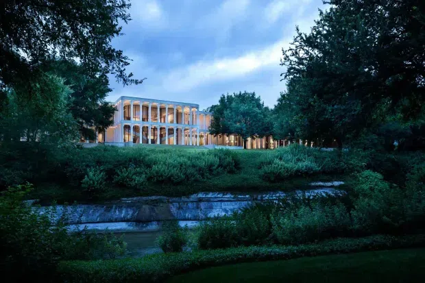 A dusk exterior view of a large contemporary multi-storey building set within a mature landscaped garden. The building features a rectangular glazed facade with repeated slender vertical columns and warm interior lighting, extending into a lower horizontal wing. Dense trees, layered planting, and a narrow landscaped water feature frame the scene.