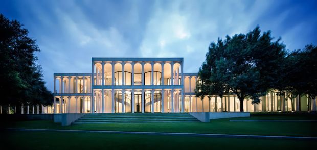 Illuminated monumental contemporary building facade at dusk, composed of long wings and a prominent central pavilion with repeated tall slender columns, large arched openings, and extensive floor-to-ceiling glazing. Broad landscaped lawn and a formal stepped entrance lead toward the building, with mature trees framing both sides.