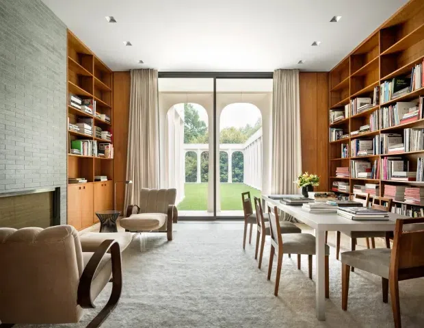 A refined, light-filled library and study with full-height warm timber bookcases and cabinetry lining both side walls. A long dining-style work table with upholstered chairs occupies the right side, while lounge chairs sit near a pale brick fireplace on the left. Floor-to-ceiling glazed doors open to a manicured garden, with tall curtains framing the view. The room has a calm, elegant, contemporary-classic character.