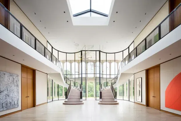 A grand, bright double-height interior atrium with symmetrical split staircases leading to an upper-level gallery. Floor-to-ceiling glazing, slender dark metal balustrades, warm timber doors and wall panels, pale flooring, and a large central skylight create an airy contemporary architectural composition. Abstract artworks are displayed along the side walls.