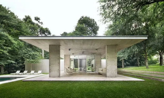 A minimalist modern pavilion set within a lush landscaped garden, featuring a broad flat roof, exposed concrete columns and ceiling, and expansive floor-to-ceiling glazing. The open-sided structure contains a refined lounge-like interior, while a swimming pool and stepped lawn landscape are visible around it.