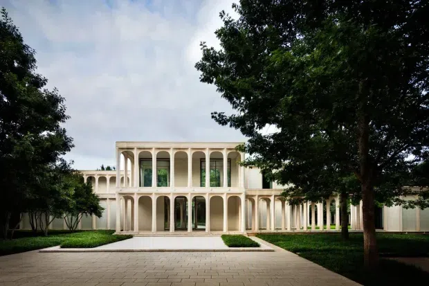 A formal two-storey cream-coloured building with a symmetrical colonnaded facade, repeated slender arches, tall glazed openings, and a central projecting pavilion. The structure is framed by mature trees and manicured lawns, with a broad paved approach in the foreground. The image presents an elegant classical-inspired exterior in soft natural light.