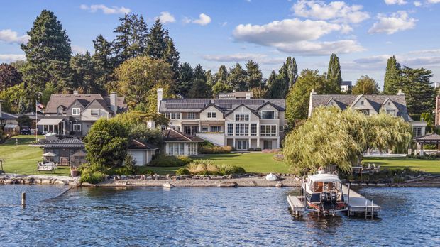 Wide exterior view of a group of waterfront residences along a calm body of water, with a prominent multi-level light-colored home, neighbouring pitched-roof houses, private lawns, mature trees, a shoreline retaining edge, and a small private dock with a boat. The scene has a clear blue sky and a suburban lakeside or riverside setting.