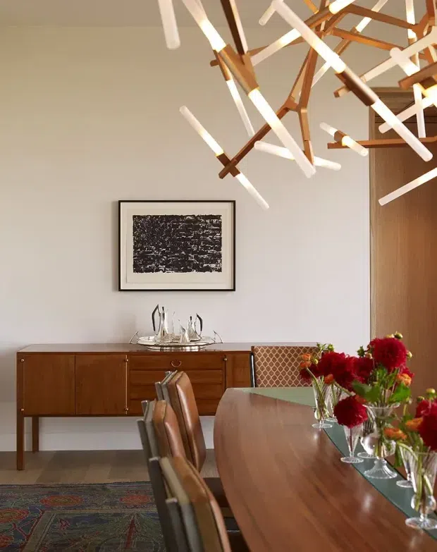 A warmly styled dining room with a long polished timber dining table, matching mid-century timber dining chairs, and a low timber sideboard against a light neutral wall. A sculptural linear pendant light with timber and white elements hangs above the table. The room includes framed monochrome artwork, glassware, a patterned rug, floral arrangements, and a textured woven chair back, creating a refined mid-century-inspired dining setting.