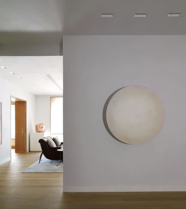 A minimalist contemporary interior view focused on a white partition wall with a large circular wall-mounted light or artwork. The foreground has pale timber flooring, white walls and ceiling with recessed linear downlights. Through the opening to the left, a refined seating area with a dark curved lounge chair, cushions, a pale rug, timber doorway and a small display plinth is visible.