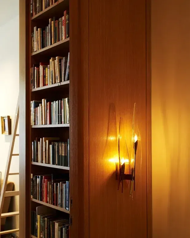 Close-up interior view of a tall, dark timber built-in bookcase filled with books beside a warm-toned timber-paneled wall. A sculptural wall sconce provides warm ambient light, while a ladder and partial shelving are visible at the left edge. The image has a rich, moody, traditional library-like character.