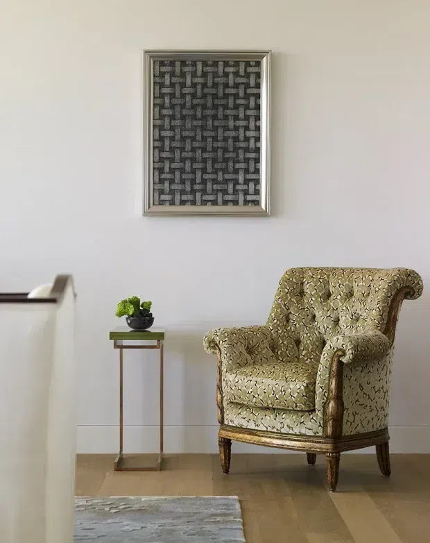 A refined interior vignette featuring a vintage upholstered wingback armchair with carved wooden legs and arms, positioned on pale timber flooring beside a slim brass-toned side table with a small potted plant. A framed geometric textile artwork hangs on the light neutral wall, while the edge of a pale console and a rug are visible in the foreground.