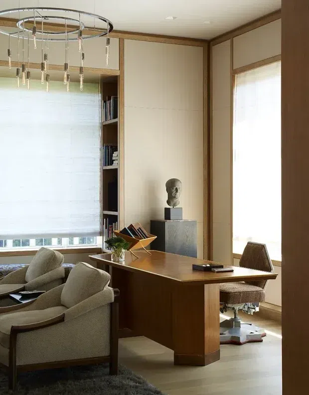 A refined wood-panelled study or home office with a substantial dark timber desk, upholstered guest chairs, built-in shelving, and a sculptural bust. Large windows are fitted with translucent roller blinds, while a circular contemporary pendant adds a focal point. The room has warm timber trim, pale wall panels, and a neutral carpet and furnishings, creating a sophisticated mid-century-inspired atmosphere.
