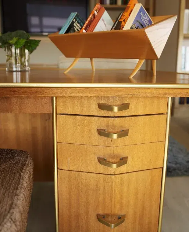 Close-up interior view of a warm mid-century-style wooden sideboard or cabinet with multiple drawers, brass-toned handles, and slender gold-colored trim. A sculptural wooden magazine rack holding books sits on top, with a blurred living area and plant in the background.
