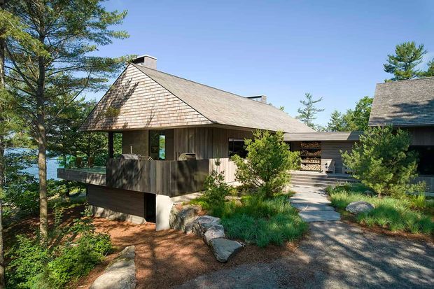 Exterior view of a low-rise timber-clad house set among mature trees and landscaped rocks, with a broad pitched gable roof clad in weathered wood shingles. The building appears elevated on a concrete base, with a covered veranda or balcony, deep eaves, dark timber walls, and a connected lower-roof wing. A glimpse of blue water is visible through the trees at left. The setting is natural, wooded, and tranquil, with gravel paths, native-style planting, and boulders around the entry.