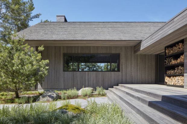Exterior view of a low-slung contemporary house with a broad shallow-pitched roof, vertical timber cladding, dark-framed horizontal window, and a raised timber deck with broad steps. A recessed opening at right contains neatly stacked firewood. The house is integrated into a natural, landscaped setting with ornamental grasses, rocks, and mature trees.