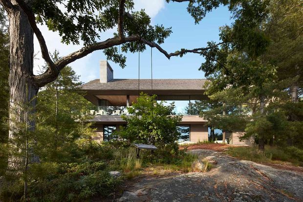 Exterior view of a contemporary, low-profile two-storey home nestled among mature trees and natural landscaping. The house features broad horizontal roof planes, extensive glazing, warm timber-toned cladding, a prominent chimney, and a suspended swing beneath a covered upper-level terrace. A rocky, informal path leads toward the entrance.